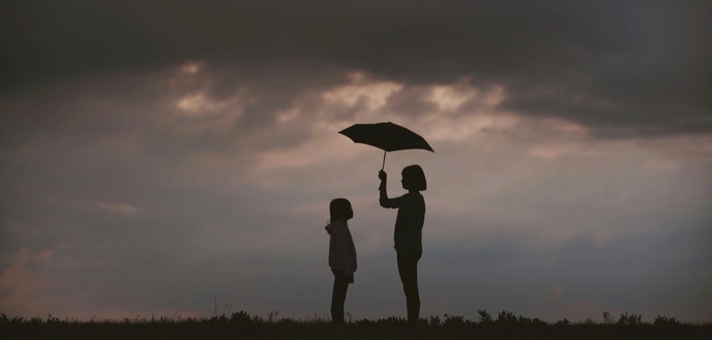 Women holding umbrella on grass field above herself and a child. Dark clouds above them.